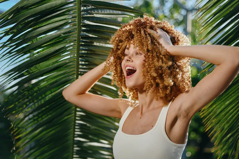 Young woman with curly hair wearing white tank top joyfully enjoying tropical palm leaves background, expressing happiness and summer vibes outdoors.