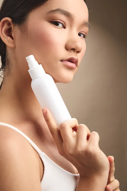 asian woman holds a white cosmetic bottle near her face in a clean studio, showcasing skincare focus, calm expression, and modern beauty routine in a minimalist setting.