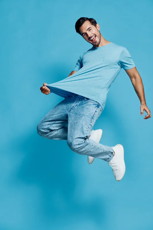 Full body joyful happy young man wearing a light blue t-shirt and denim jeans jumps in the air, expressing excitement against a bright blue background. Lifestyle concept.