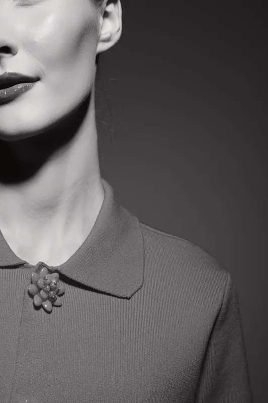 Close-up of a white woman with pale skin wearing a collared shirt with a decorative brooch, captured in moody black and white lighting. Concept of minimalism and fashion.