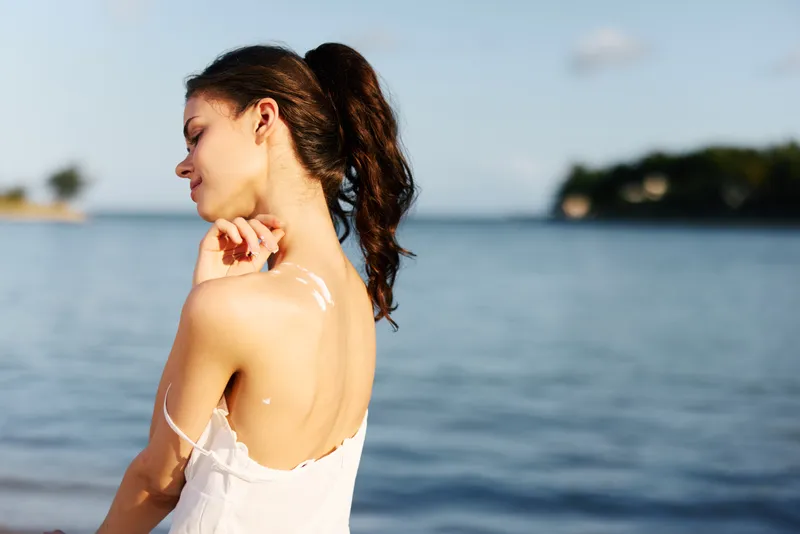 Young woman applying sunscreen on her shoulder by the sea on a sunny day with calm water and distant islands.