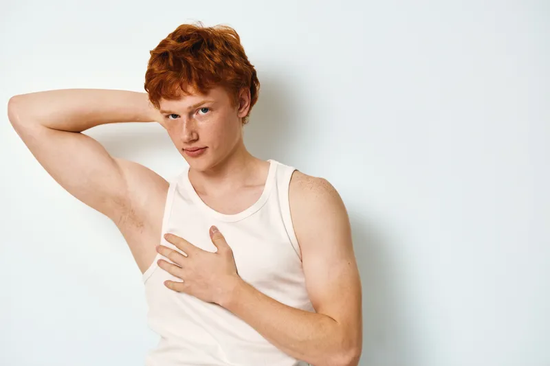 Young man with red hair in white tank top posing with hand under arm and other on chest standing against white background closeup portrait studio shot expression.