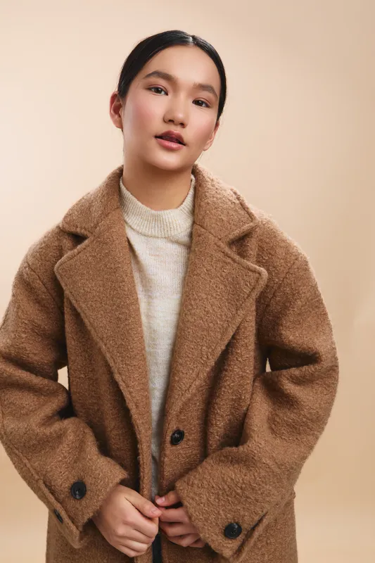 Asian woman stands in a neutral studio, wearing a cozy oversized brown coat over a light sweater, exuding calm confidence and warmth and approachable vibe.