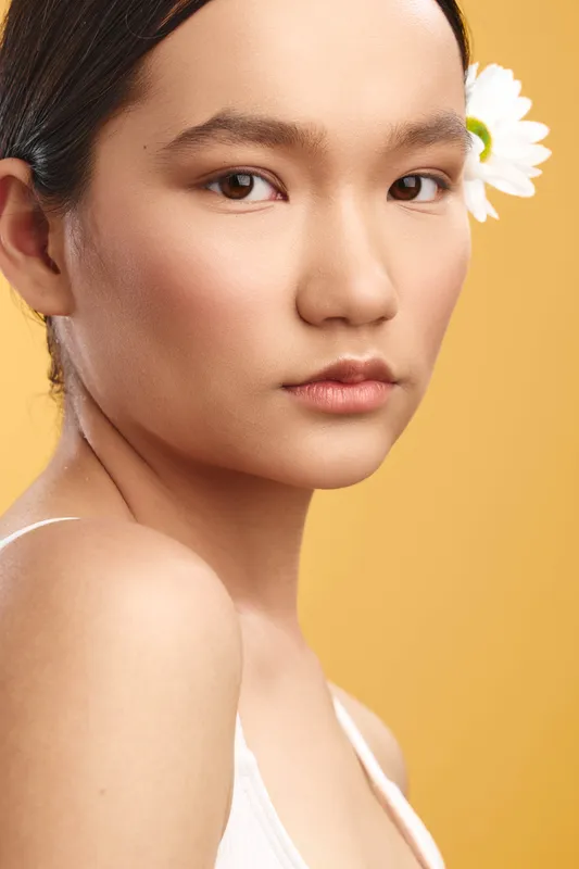 Portrait of an asian woman with a flower behind her ear, set against a warm yellow backdrop. Soft lighting highlights smooth skin, conveying calm beauty and natural elegance.
