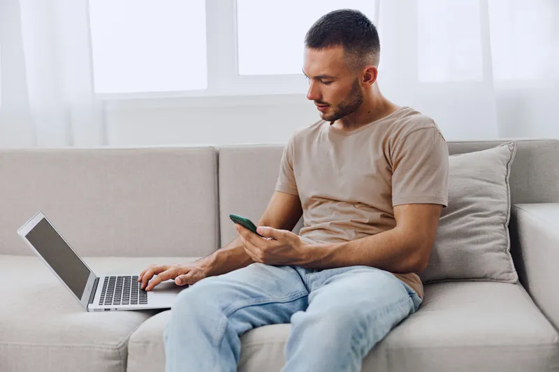 Focused man working on laptop while using smartphone. Modern workspace with natural light, reflecting productivity and digital connectivity in 2025.