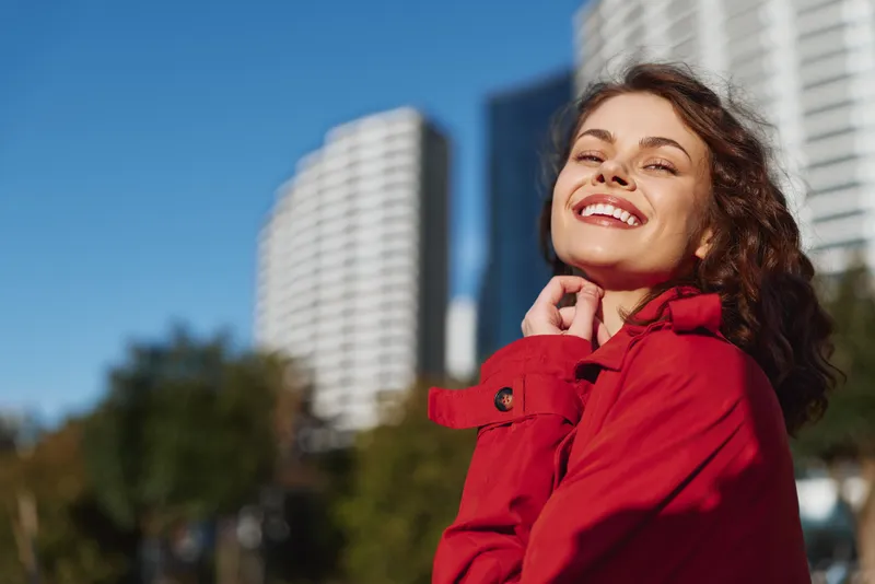 Smiling young woman in a red coat stands on a sunny city street, her confident expression highlighted by tall buildings rising behind her in a vibrant urban portrait.