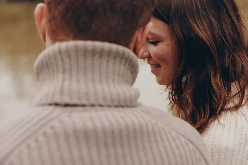 Candid lifestyle moment showing a couple sharing a gentle smile and close connection outdoors, wearing cozy sweaters, intimate warmth, natural light, calm mood, affectionate partnership