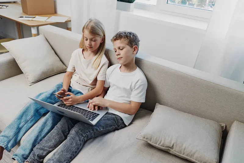 Two children sitting on a sofa, a boy using a laptop and a girl holding a smartphone, enjoying digital devices together in a bright modern living room environment.