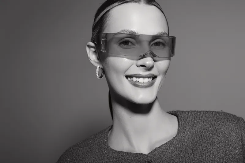 Smiling Caucasian woman with fair skin wearing transparent glasses and textured sweater, posing in studio with soft lighting and neutral background.