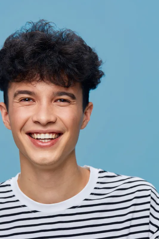 smiling man with curly hair wearing striped shirt on blue background, joyful expression, casual style, close-up portrait, young adult male happiness