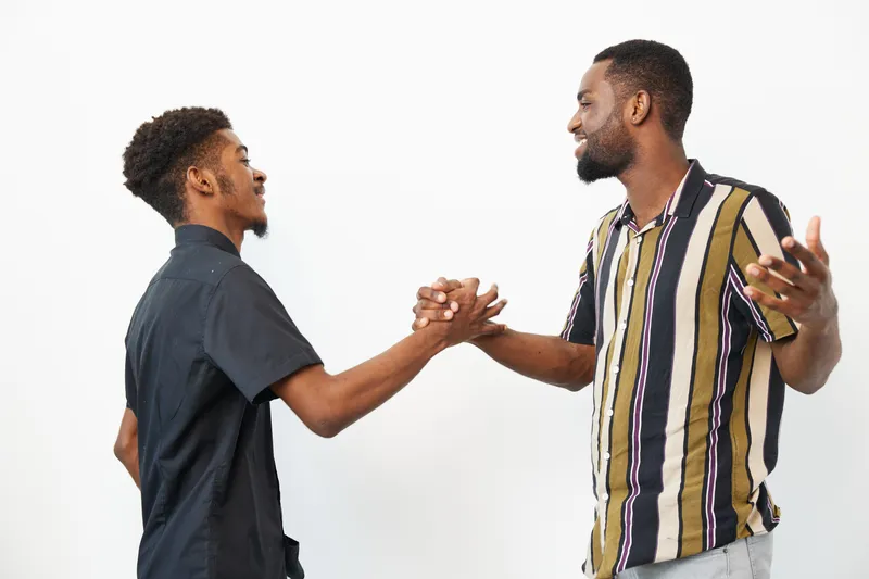 Two joyful African American men greeting each other with smiles and handshakes against a bright background. Celebrating friendship and connection in a lively atmosphere.
