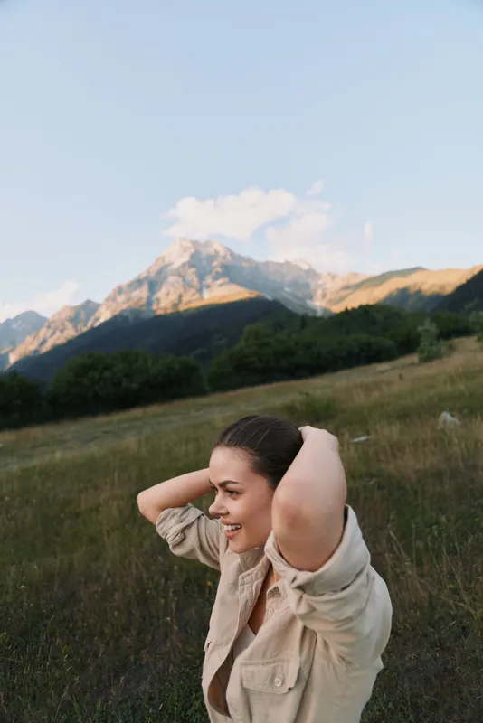 A woman enjoys an outdoor moment in a grassy field with distant mountains, light wind brushing her hair. A candid lifestyle image capturing natural joy and serene nature.