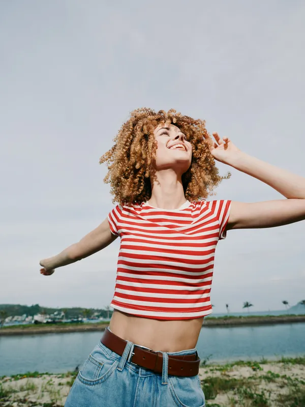 Young woman with curly hair wearing a red and white striped crop top and denim jeans, smiling and dancing outdoors by a calm waterfront on a sunny day.