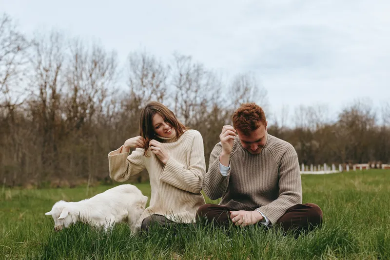 Couple sitting in a grassy field with a playful goat kid nearby, sharing a relaxed moment. Cozy sweaters, gentle light, and rural lifestyle create a warm, wholesome mood.