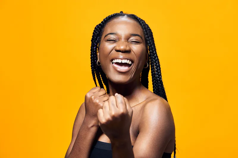 Joyful young woman celebrating success with braided hair against a vibrant yellow background, expressing happiness and optimism in her stylish black outfit