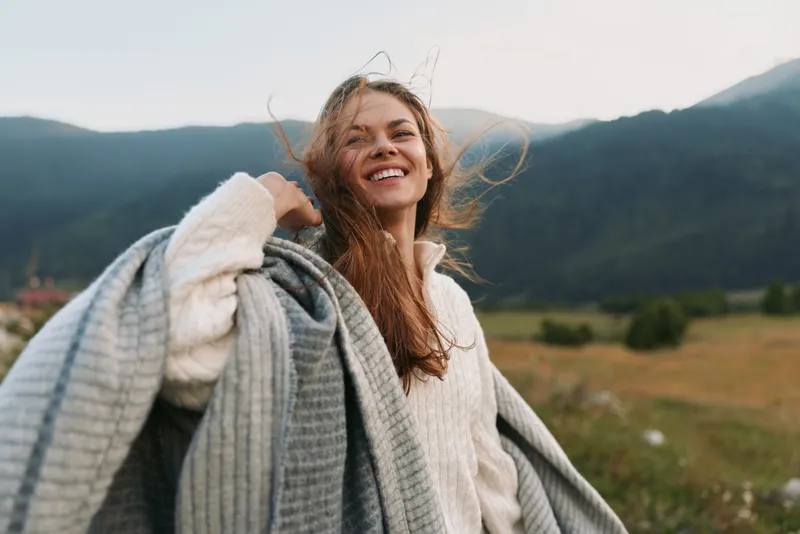 Joyful woman in cozy knitwear outdoors, standing in a grassy valley with mountains in the background, enjoying fresh air and a relaxed lifestyle moment, smiling.