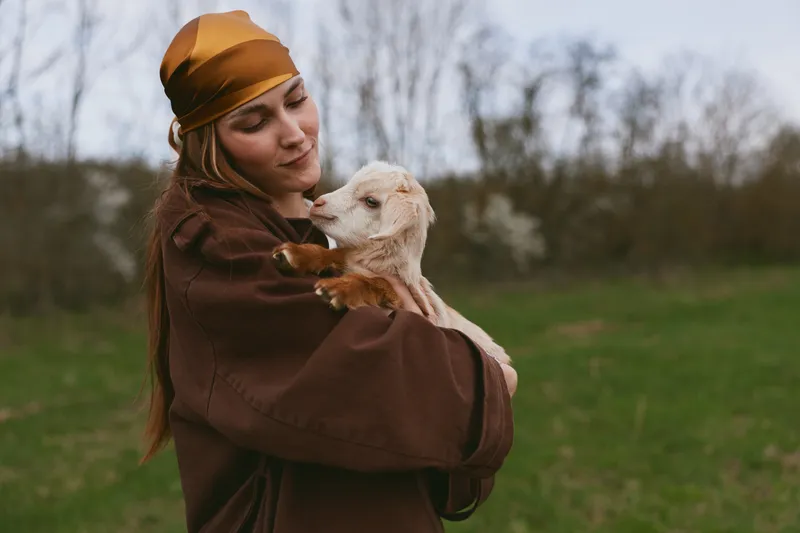 Young woman in brown jacket and yellow headscarf gently holds a small white baby goat outdoors in a green field during early spring.