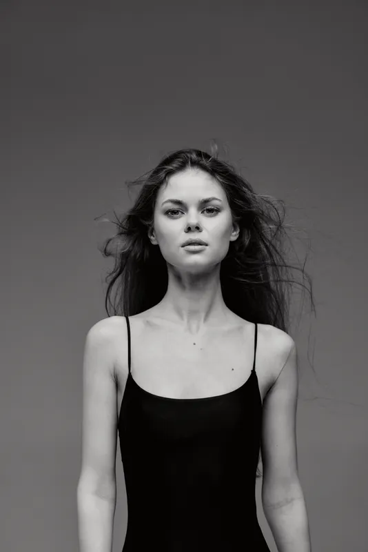Black and white portrait of a young woman with loose hair wearing a black sleeveless top against a plain background.