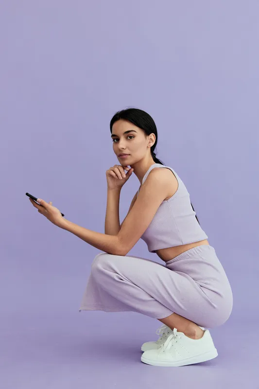 Young woman crouching in casual lilac outfit and white sneakers, holding a smartphone with thoughtful expression against plain purple background.