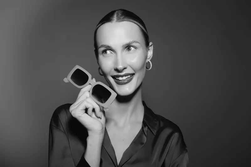 Portrait of a smiling Caucasian woman with light skin holding sunglasses near her face. She wears hoop earrings and a silky blouse against a plain studio background. Concept for fashion and style.