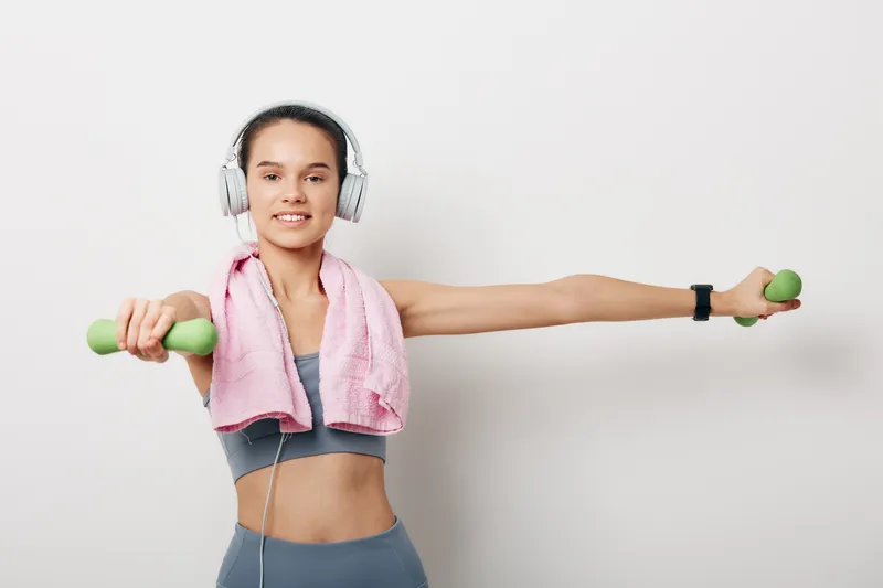 Woman in athletic wear with headphones and a towel, holding small dumbbells in a wide arm stretch, ready for a high-energy fitness workout at home.