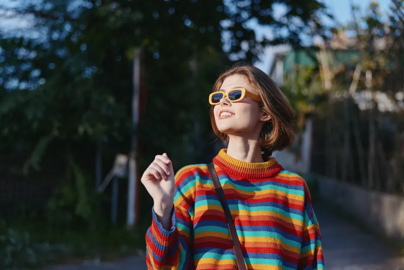 Young woman in her 20s smiling and looking up outdoors, wearing a striped sweater and yellow sunglasses with short hair and crossbody bag, sunny lifestyle portrait.