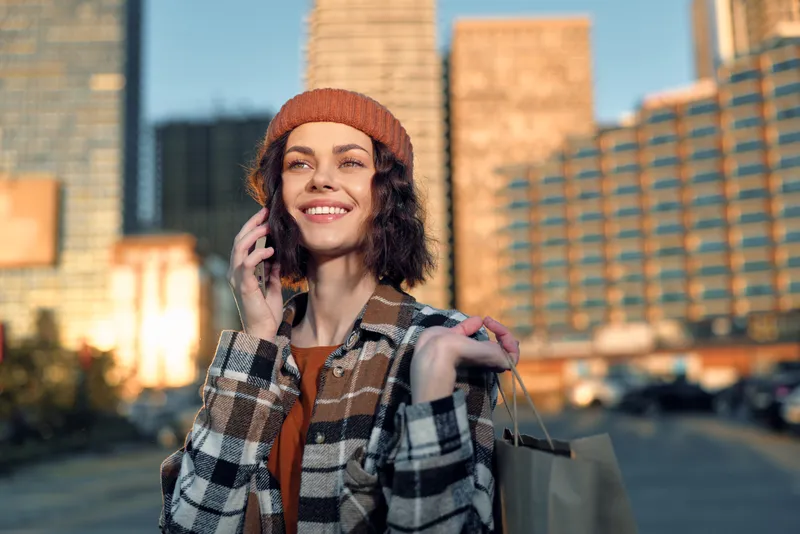 Woman on phone with shopping bags, smile framed against urban city skyline at golden hour, candid lifestyle portrait conveying authenticity, mindful living and emotional storytelling.