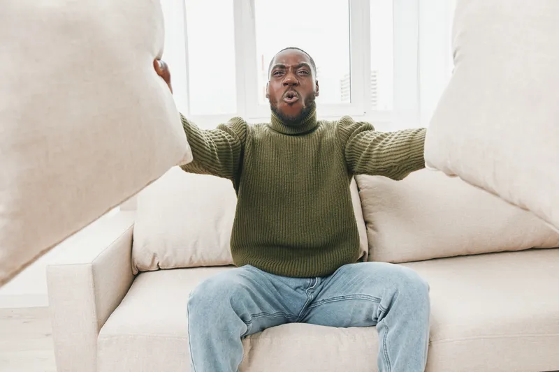 Young African American man expressing frustration on a couch while holding pillows, wearing a green sweater, casual jeans, in a bright modern interior