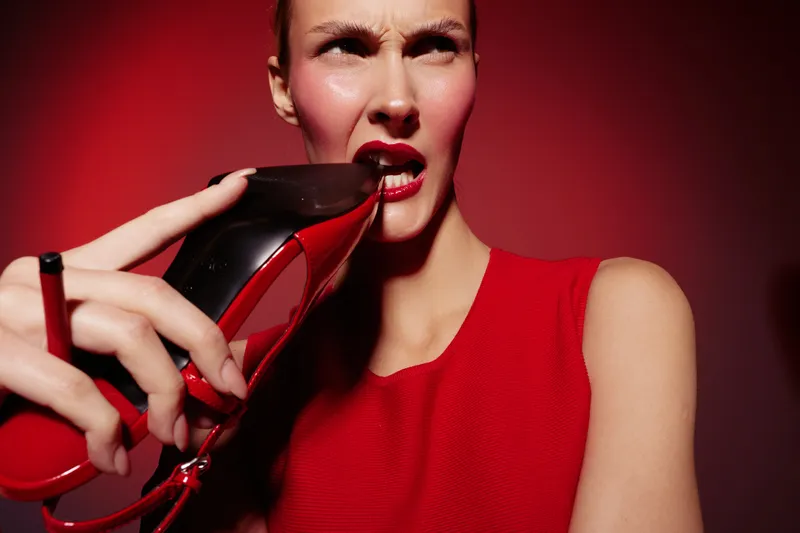 Woman in red dress bites red high heel shoe with intense expression on red background. Concept of fashion, attitude, and bold style in studio lighting.