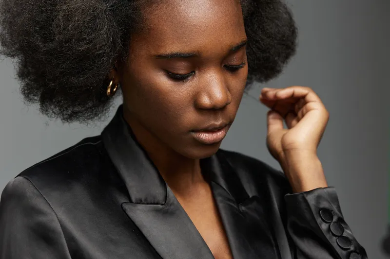 Portrait of a young Black woman wearing a black satin blazer, displaying introspective emotions, against a muted gray background, emphasizing elegance and style
