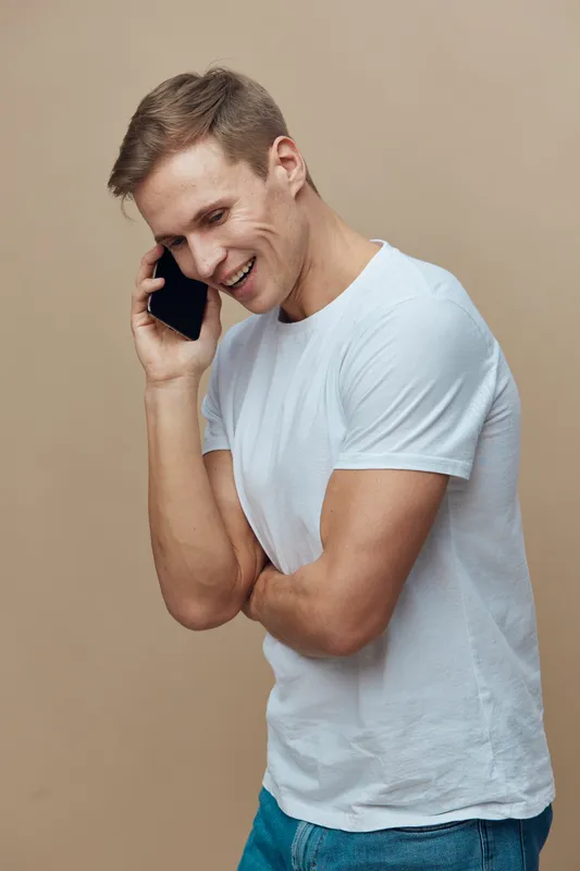 Smiling man talking on mobile phone, casual white t-shirt, young adult, cheerful expression, beige background, lifestyle shot, happy mood, communication concept, modern fashion