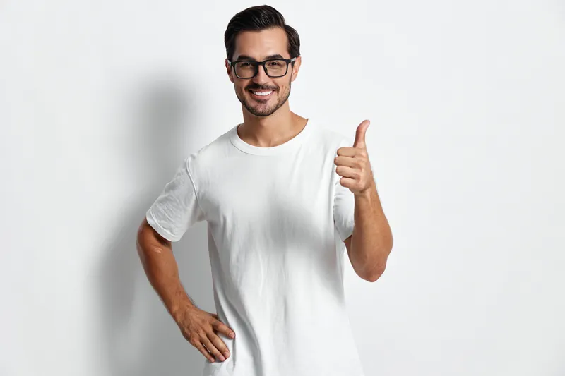 Smiling man wearing glasses and a white t-shirt shows a thumbs up gesture, standing against a plain solid background. Positive attitude and cheerful expression.