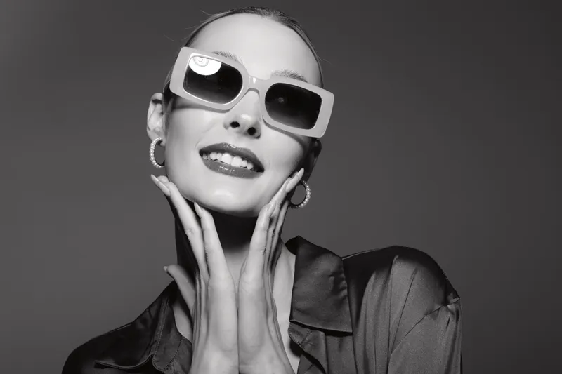 A smiling young white woman with pale skin wearing large sunglasses and hoop earrings poses with hands near face in a stylish black outfit. Concept of fashion and glamour.