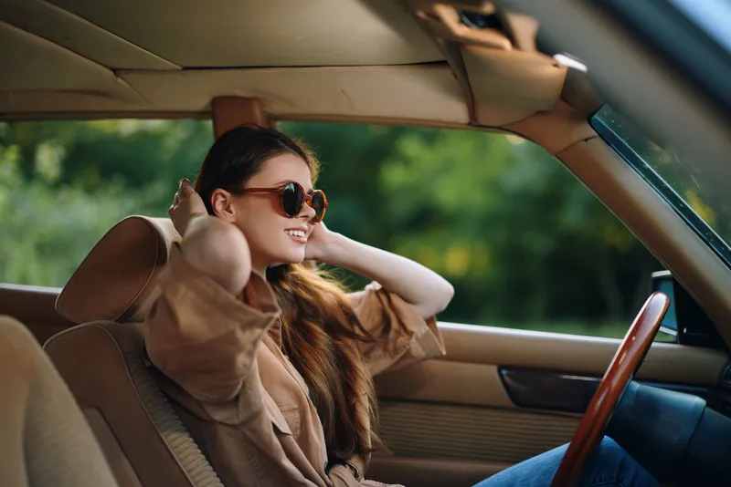 Young woman enjoying the sun in a car, wearing sunglasses and smiling, with a lush green background, capturing a feeling of freedom and joy during summer.