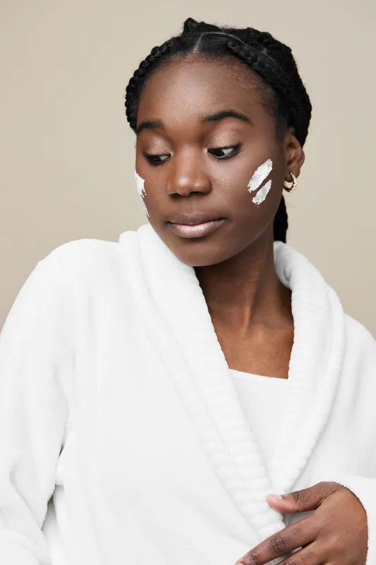 A young woman of African descent in a white robe applying skincare products on her face, with a serene expression, against a neutral background