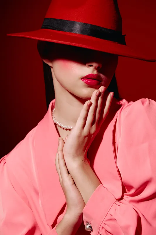 Stylish woman in a bold red hat and lips poses with hands near face, wearing a pink blouse and pearl necklace. Creative beauty fashion portrait with strong red tones and dramatic shadows.