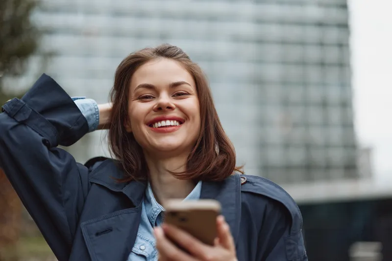 A smiling woman outdoors with a smartphone in hand, wearing a casual jacket. Concept of everyday confidence, digital communication, and upbeat urban lifestyle.