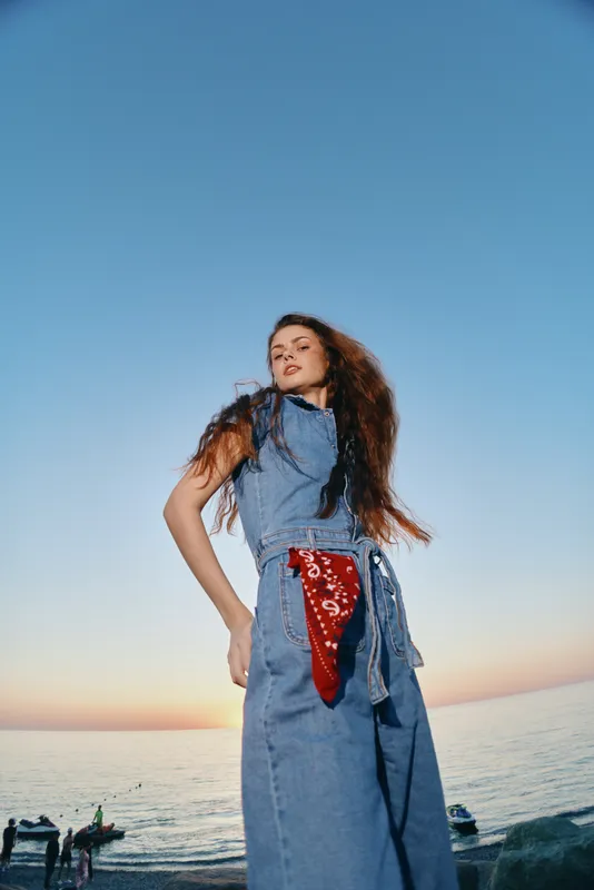 Lifestyle woman in modern boho-western denim street style poses confidently against a serene ocean sunset, showcasing vibrant film color and relaxed summer fashion.