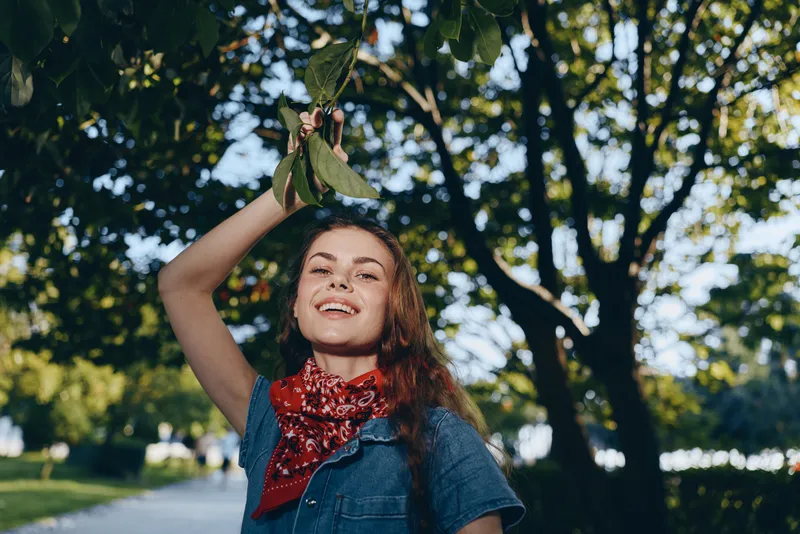 Lifestyle woman in modern boho-western denim street style smiling outdoors under green trees with film color tones, enjoying sunny day and casual fashion vibes.