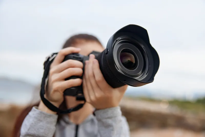 Young woman with long brown hair holding a DSLR camera, capturing outdoor photography with enthusiasm and creativity, in a scenic natural background