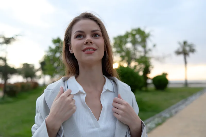 Young woman outdoors smiling confidently in casual white shirt with soft natural light and green park background, lifestyle, spring, optimism, portrait