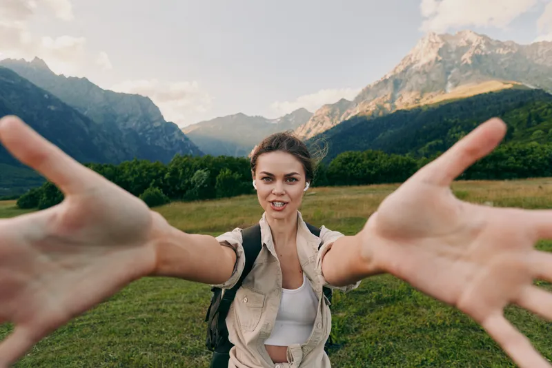 Young woman with a backpack reaches out towards the camera while hiking in a mountainous landscape under a partly cloudy sky.