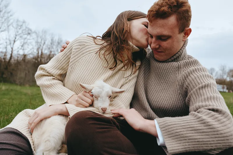 Smiling couple with a white goat kid cuddling outdoors in knit sweaters. Close-up on a grassy field under an overcast sky. Lifestyle scene of affection, nature, rustic comfort, and shared tenderness.