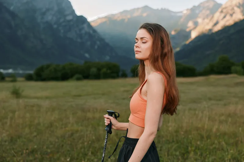 A serene young woman stands in a scenic valley, holding a trekking pole. She wears a coral athletic top and dark pants, eyes closed, savoring fresh air, calm focus, and a peaceful outdoor moment.