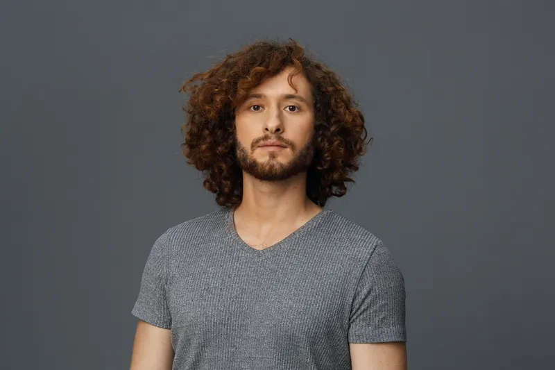 Young man with curly hair in a casual gray t shirt, expressing confidence and calmness against a muted background, showcasing modern fashion and personality