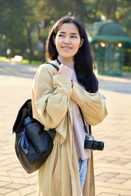 Woman wearing a light coat with backpack and camera in park, enjoying outdoor stroll, sunlight, casual style and candid moment captured during daytime in open space