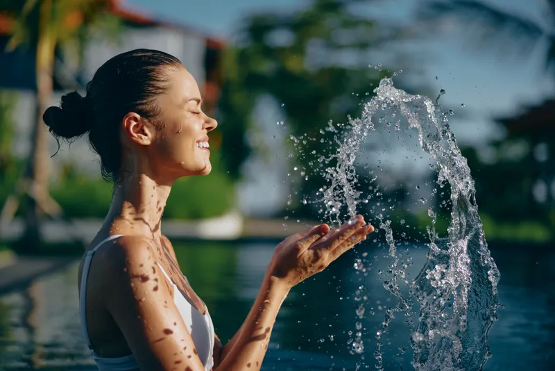 A smiling woman enjoys a refreshing water spray beside an outdoor pool in bright sunshine, capturing a joyful, carefree summer moment and a relaxed lifestyle, with warm light, playful droplets, and