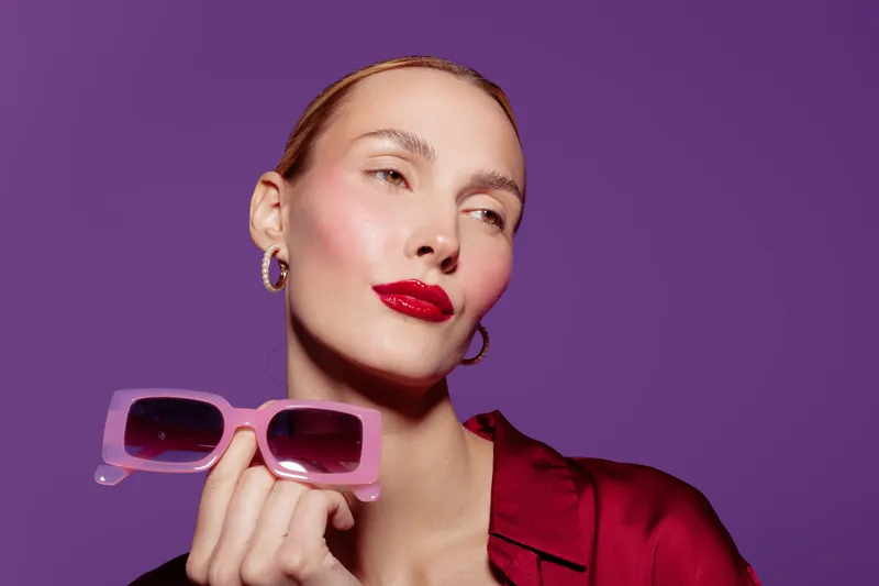 Stylish woman with red lipstick holds pink sunglasses against purple background. Fashion portrait with confident expression and trendy accessories in studio lighting.