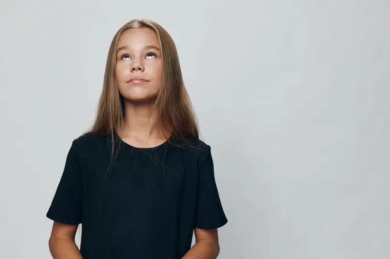 Girl with long straight hair and black shirt looking up thoughtfully on gray background. Child with expression of curiosity and casual outfit in studio with soft lighting.