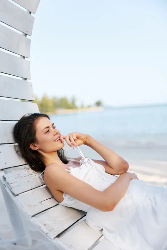 A smiling woman rests on a sunlit beach, leaning against a white wooden structure, wearing a light dress, enjoying a peaceful seaside moment and gentle breeze.